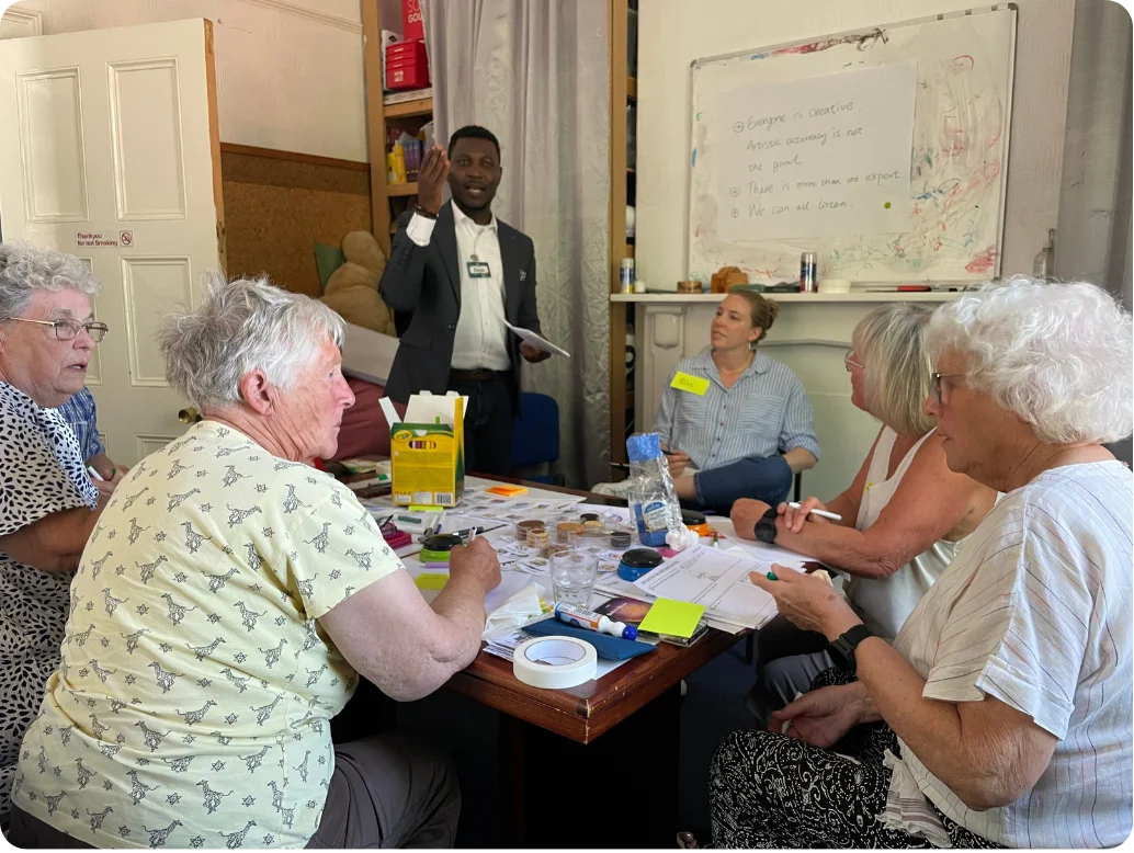 Dayo facilitating a co-design workshop with older adults at MHA (Methodist Homes Housing Association), Leeds. The whiteboard reads: Everyone is creative, Artistic accuracy is not the goal, There is more than one expert, We can all learn.